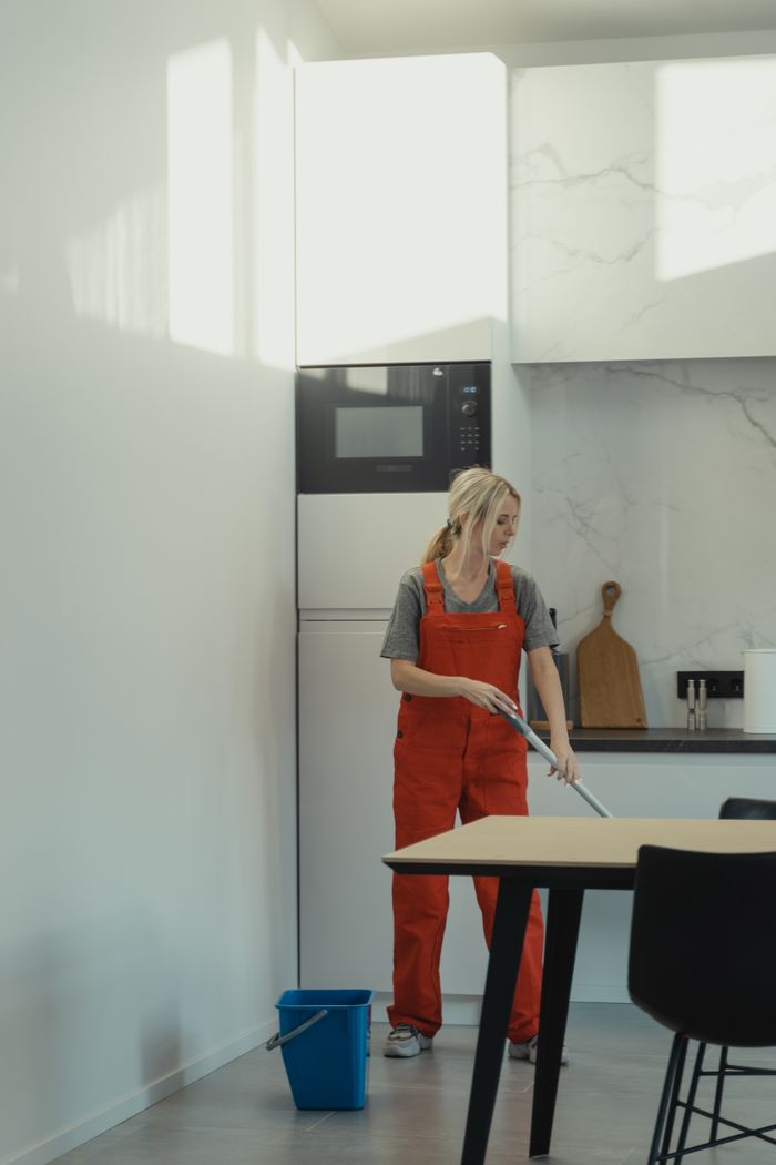 Une femme en salopette rouge nettoie le sol d’une cuisine sous une lumière vive de jour, illustrant les tâches ménagères.